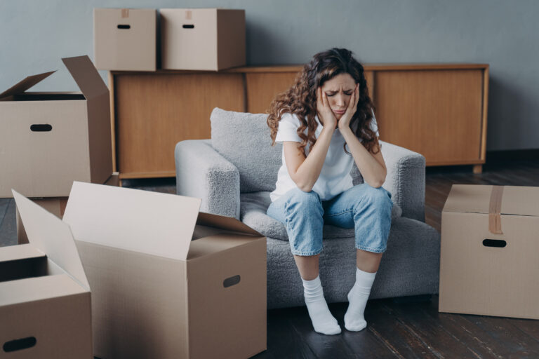 Exhausted upset woman tenant sitting on armchair in empty room near cardboard boxes feels unmotivated to pack her things. Financial problem, eviction, relocation day concept.