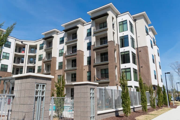 Modern multi-story apartment building with balconies, white and brown exterior, surrounded by greenery under a clear blue sky. Experience peace of mind without the hassle of problem tenants disrupting this serene environment.