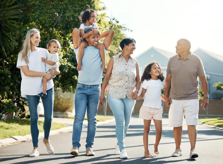 A diverse multi-generational family of six, including two children, is walking down a sunny suburban street, chatting and smiling, while discussing the importance of family wills and trusts in securing their future.