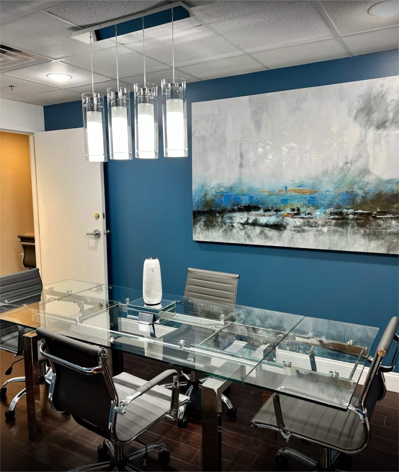 A conference room with a glass table, gray chairs, a large abstract painting, and modern pendant lights.